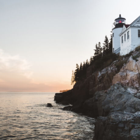 White house next to trees and a rocky cliff above the ocean with a sunset in the background