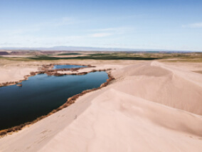 A small body of water surrounded by tan sand dunes with a light blue sky